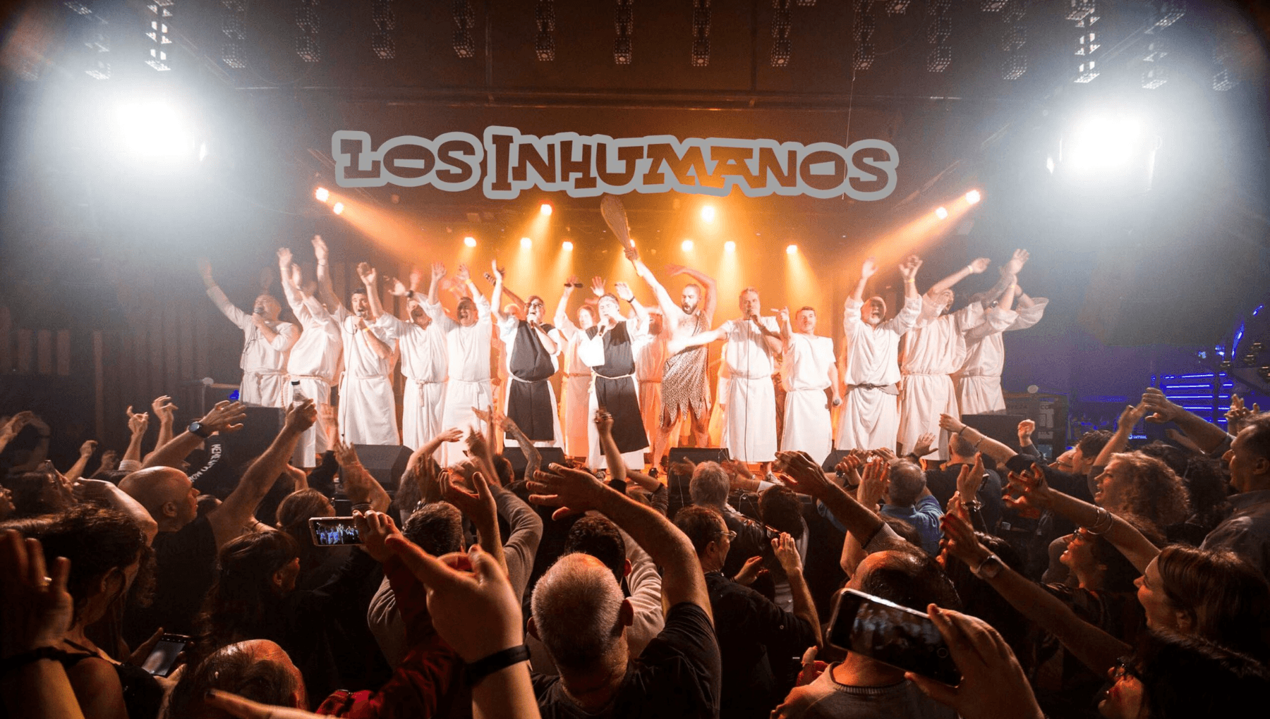 Los Inhumanos en directo, con toda la formación sobre el escenario vestida con túnicas blancas, ante un público entregado con los brazos en alto.